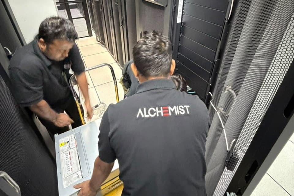 Two Alchemist technicians in branded polo shirts lifting a server chassis into a rack.