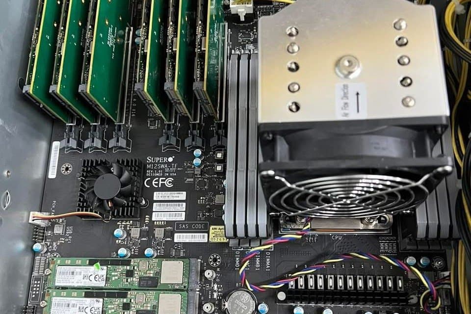 An open server chassis showing six add-in cards in PCIe slots beside a large air cooler and a bank of M.2 drives.